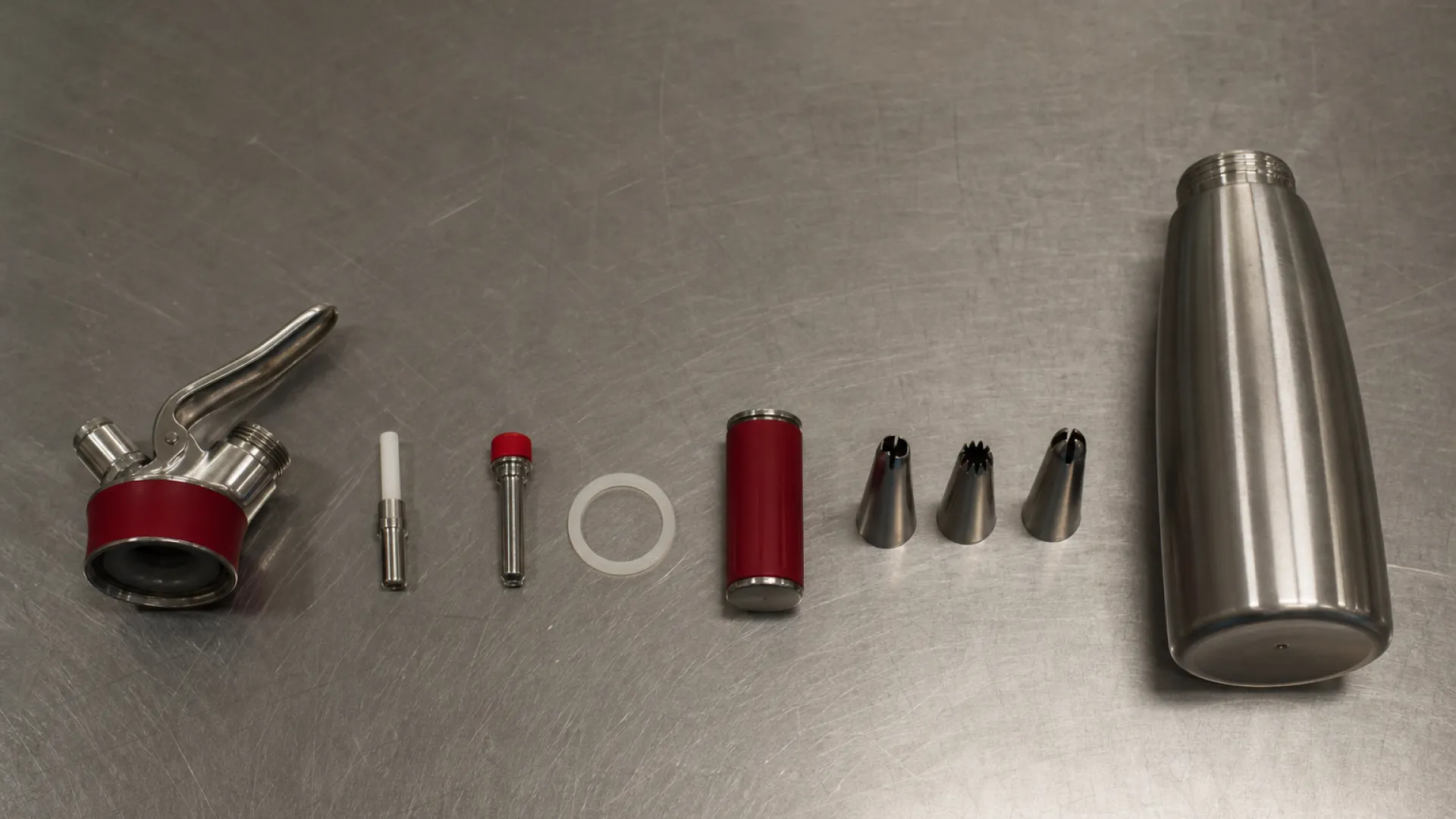 Exploded diagram of whipped cream dispenser parts: head, valve, gasket, charger holder, nozzle, canister
