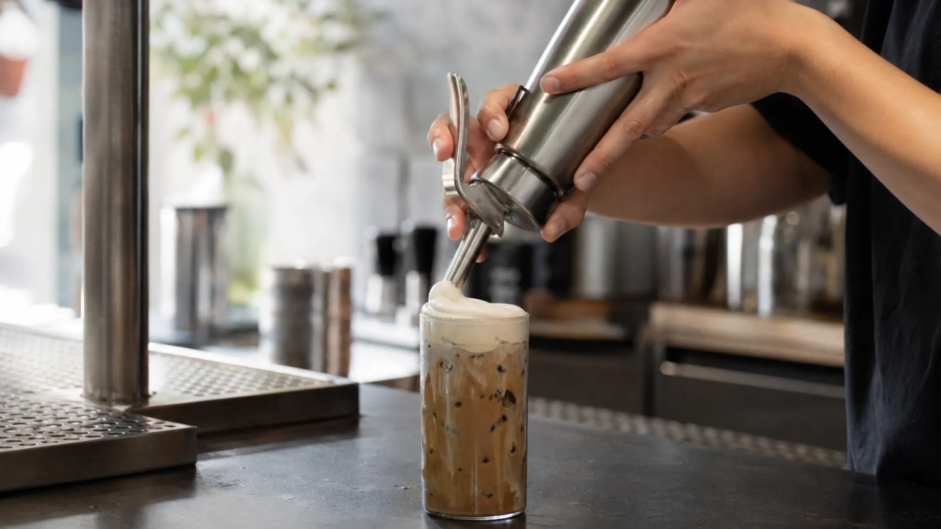 Barista topping an iced coffee with cold foam dispensed from a stainless steel cream whipper in a specialty coffee shop