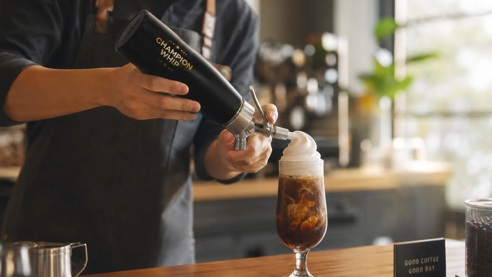 Cream charger uses in cafés: cold foam dispensed from a stainless steel cream whipper onto an iced coffee