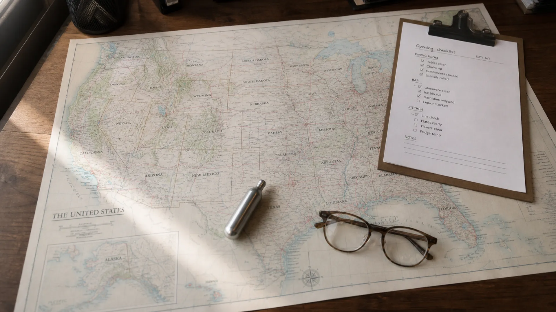 Map of the United States on a restaurant office desk with a whipped cream charger, compliance checklist, and reading glasses